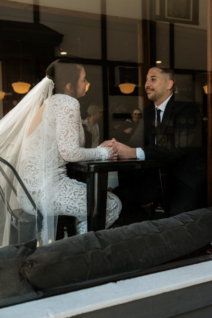 Bride and groom hold hands in coffee shop and photo taken from outside of the store.