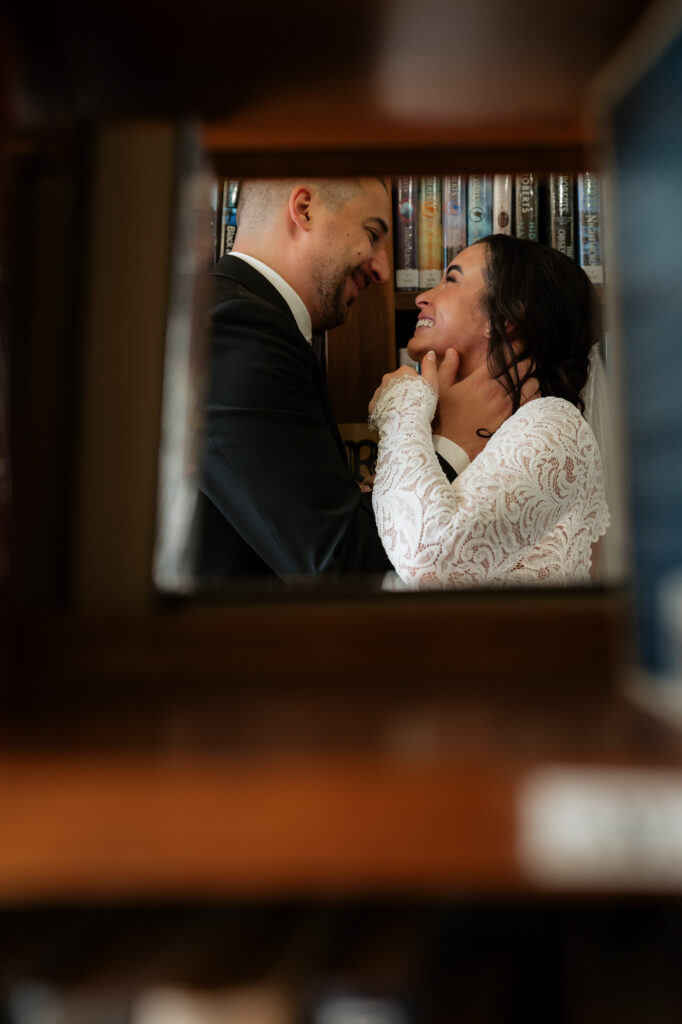 A couple is seen through a hole in a book shelf laughing at each other romantically