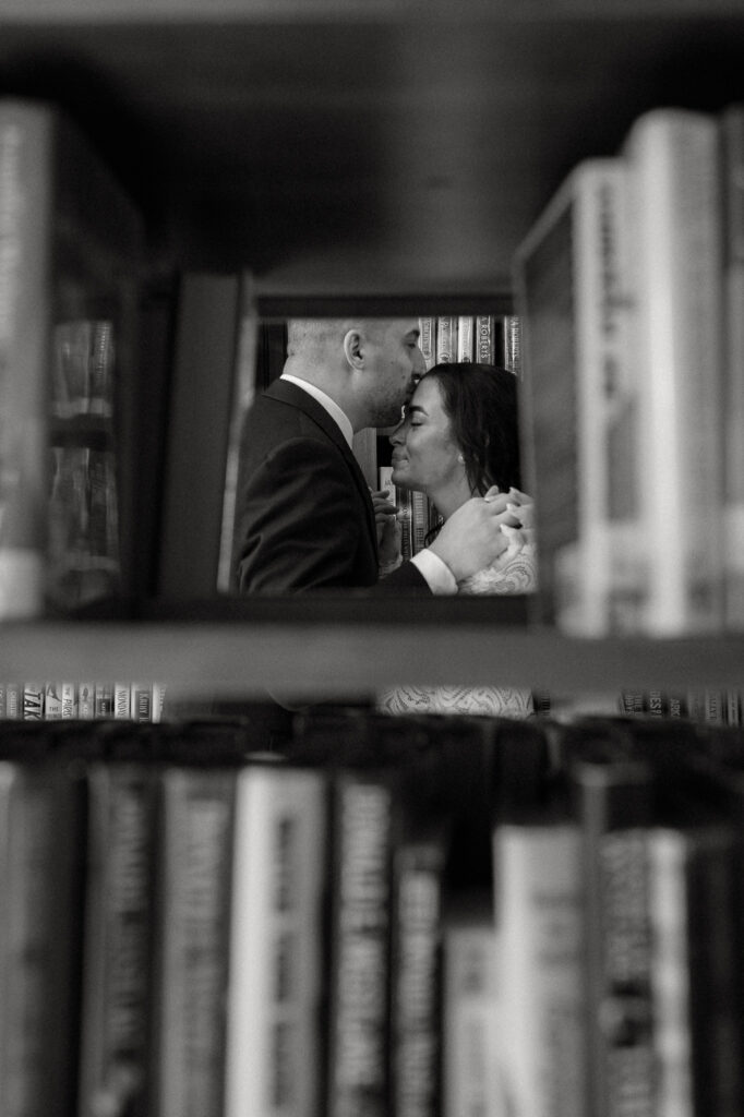 A couple is seen through a hole in a book shelf embracing where the groom kisses the brides forhead.