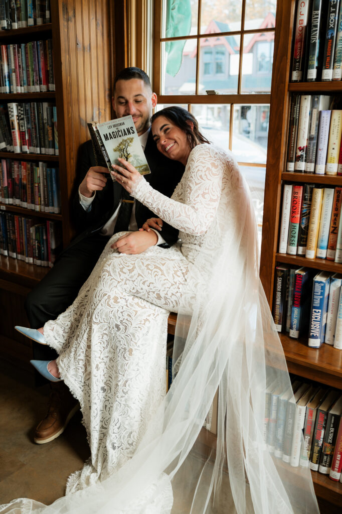 A couple is cuddled up in a library window reading books together on their wedding day.