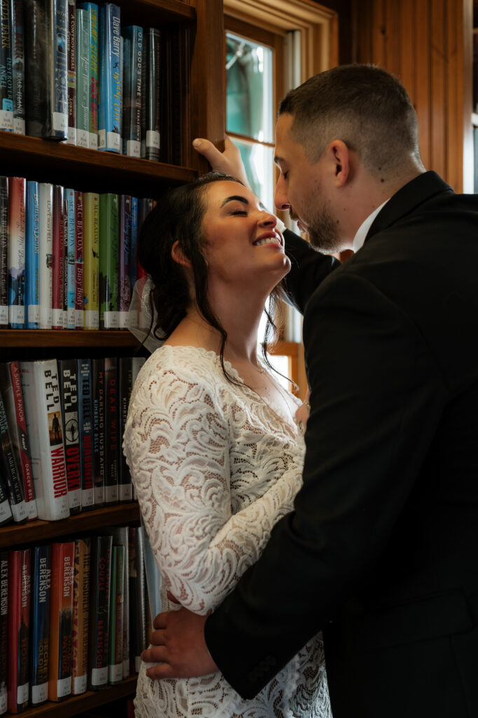 A bride is leaned up against a bookshelf by the groom and she is grabbing his suit jacket and pulling him in for a kiss.