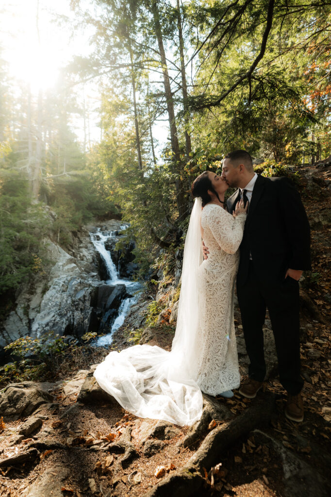 A bride kisses the groom with sunlight peeking through the forest trees and a double tiered waterfall flows behind them in the Adirondacks.