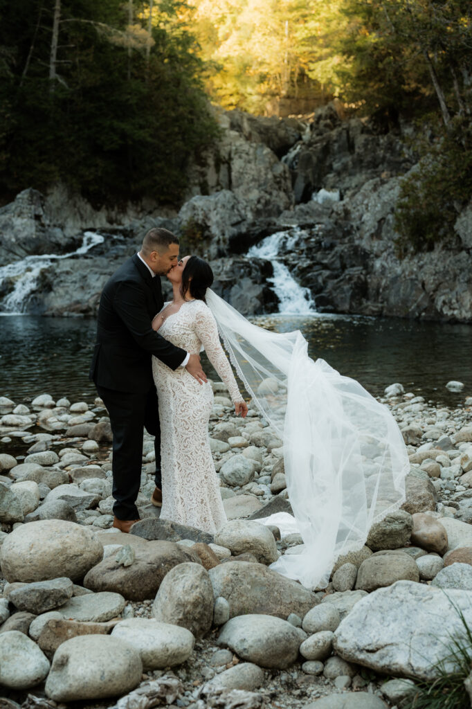 Bride and groom kiss while a veil is flowing in the wind at the base of a waterfall in the Adirondacks.
