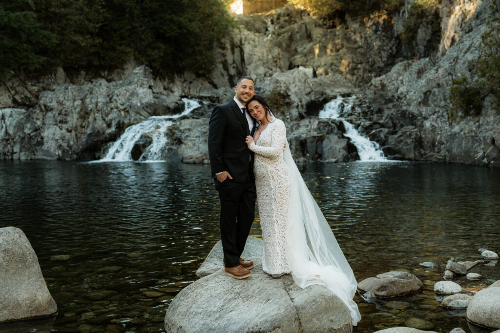 A bride and groom stand on a rock in front of a waterfall in keene valley.