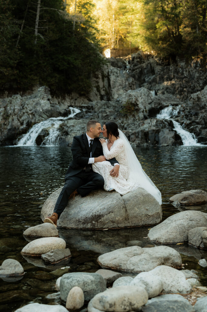 A bride and groom sit on a rock in front of a waterfall in Elizabethtown New York.