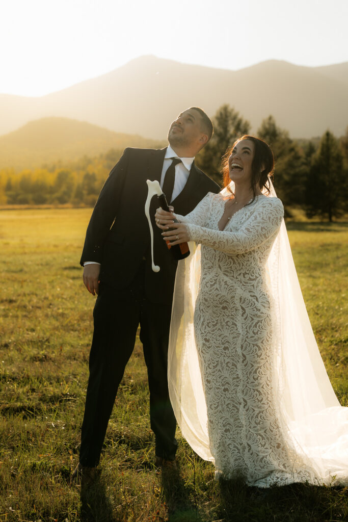 A bride and groom pop champagne in a field in the adirondacks with a mountain backdrop at sunset.