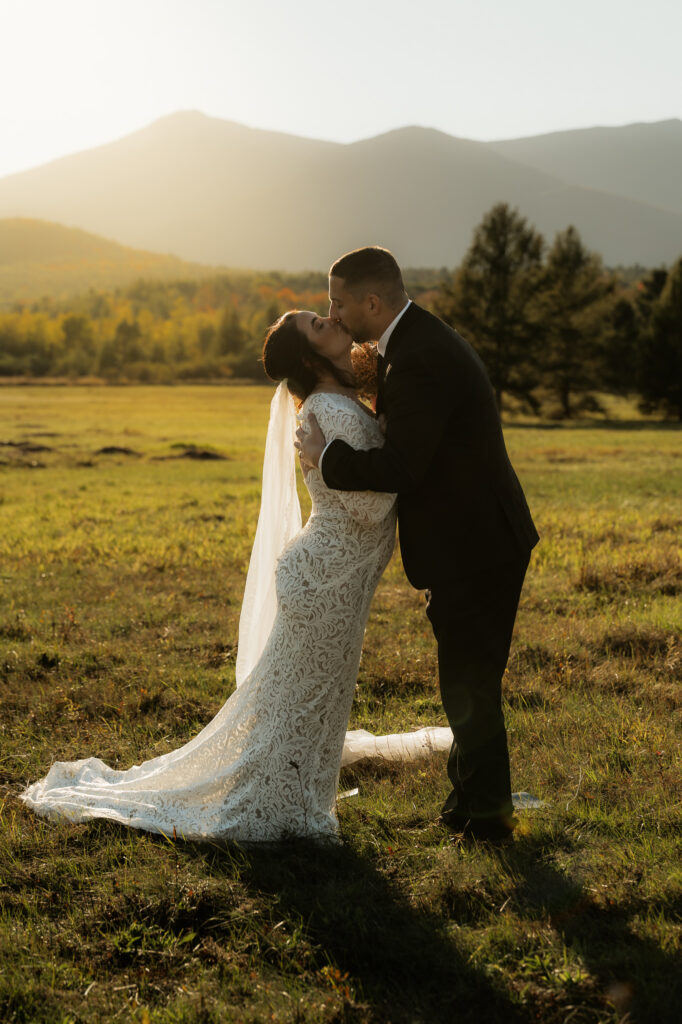 A couple kisses in a field in the adirondacks with a mountain backdrop at sunset.