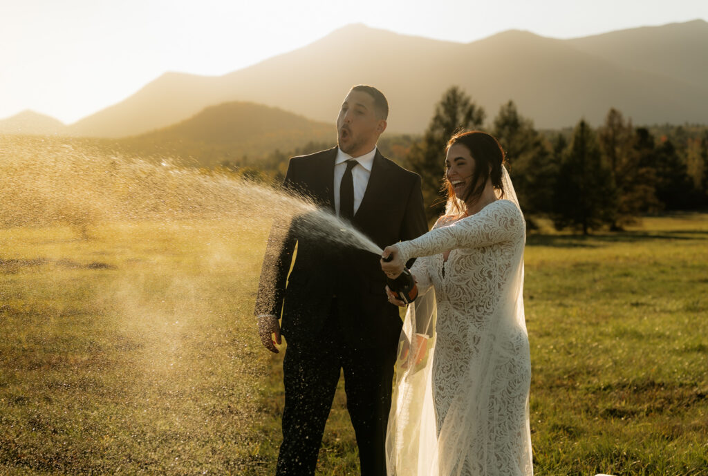 A bride and groom pop champagne in a field in the adirondacks with a mountain backdrop at sunset.