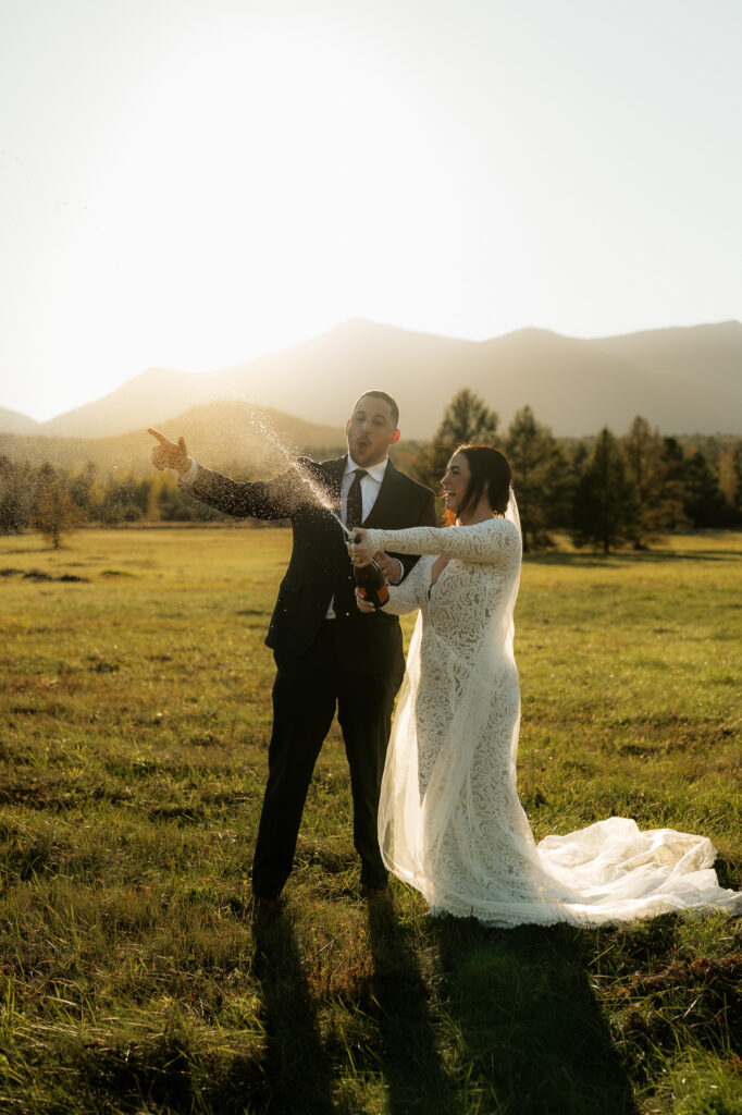 A bride and groom pop champagne in a field in the adirondacks with a mountain backdrop at sunset.