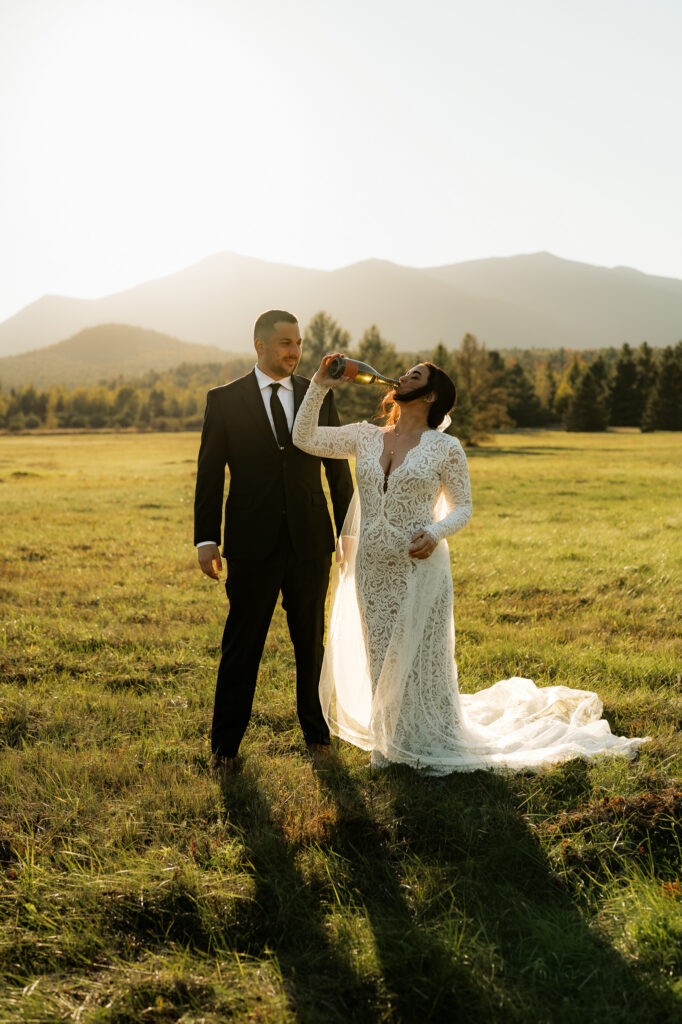 A bride and groom pop champagne in a field in the adirondacks with a mountain backdrop at sunset.