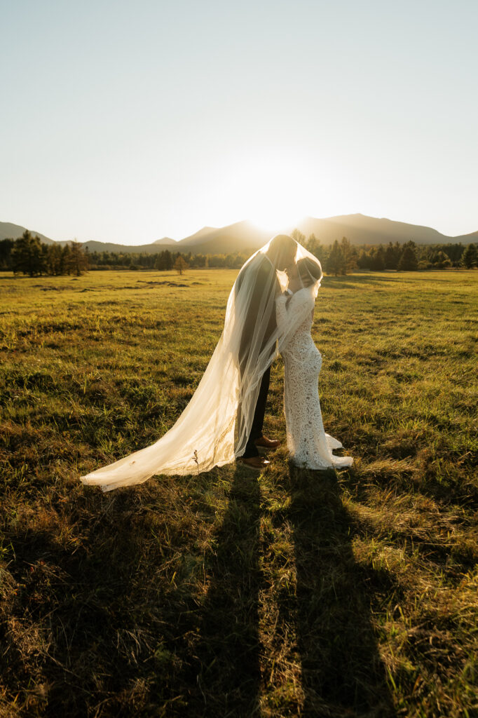A couple kisses at sunset with a veil covering the both of them in a field in the adirondacks.