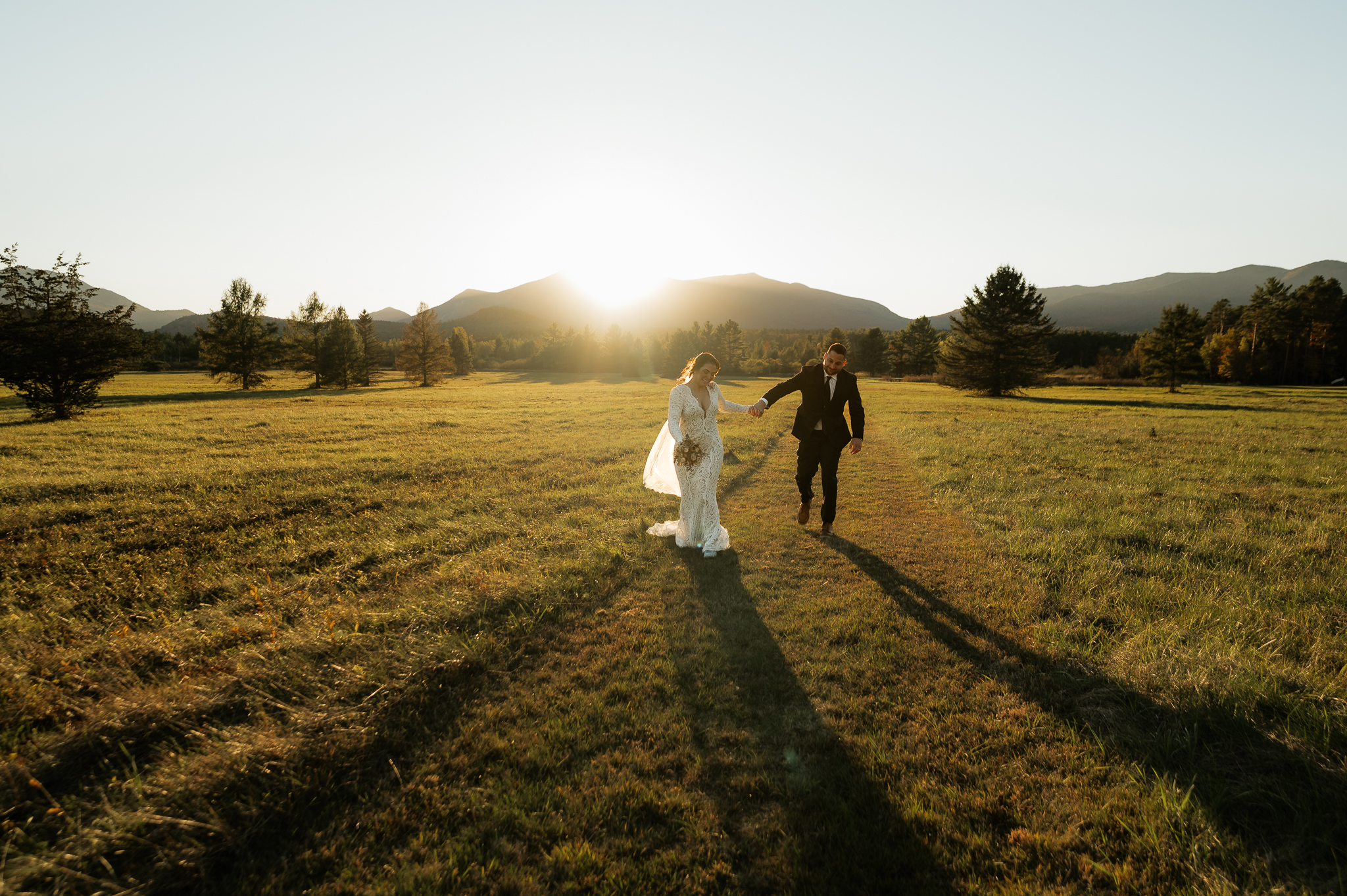 A couple frolics through an Adirondack field at sunset with a mountain backdrop.