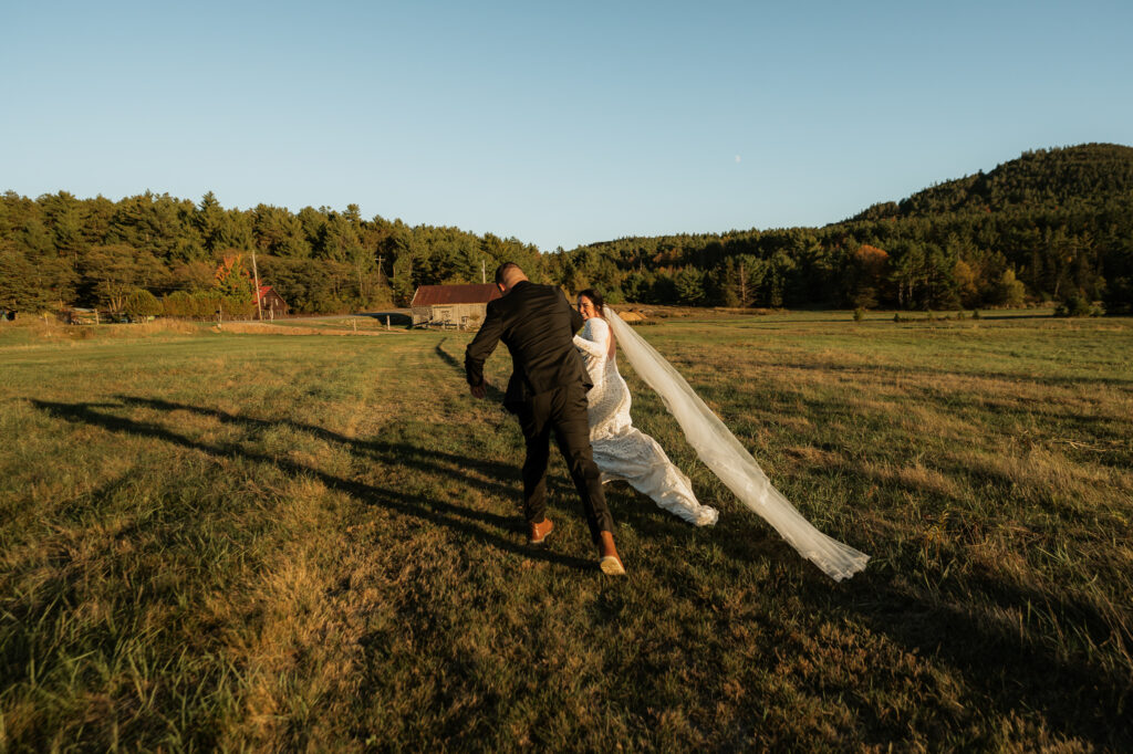A couple frolics through an Adirondack field at sunset with a mountain backdrop.
