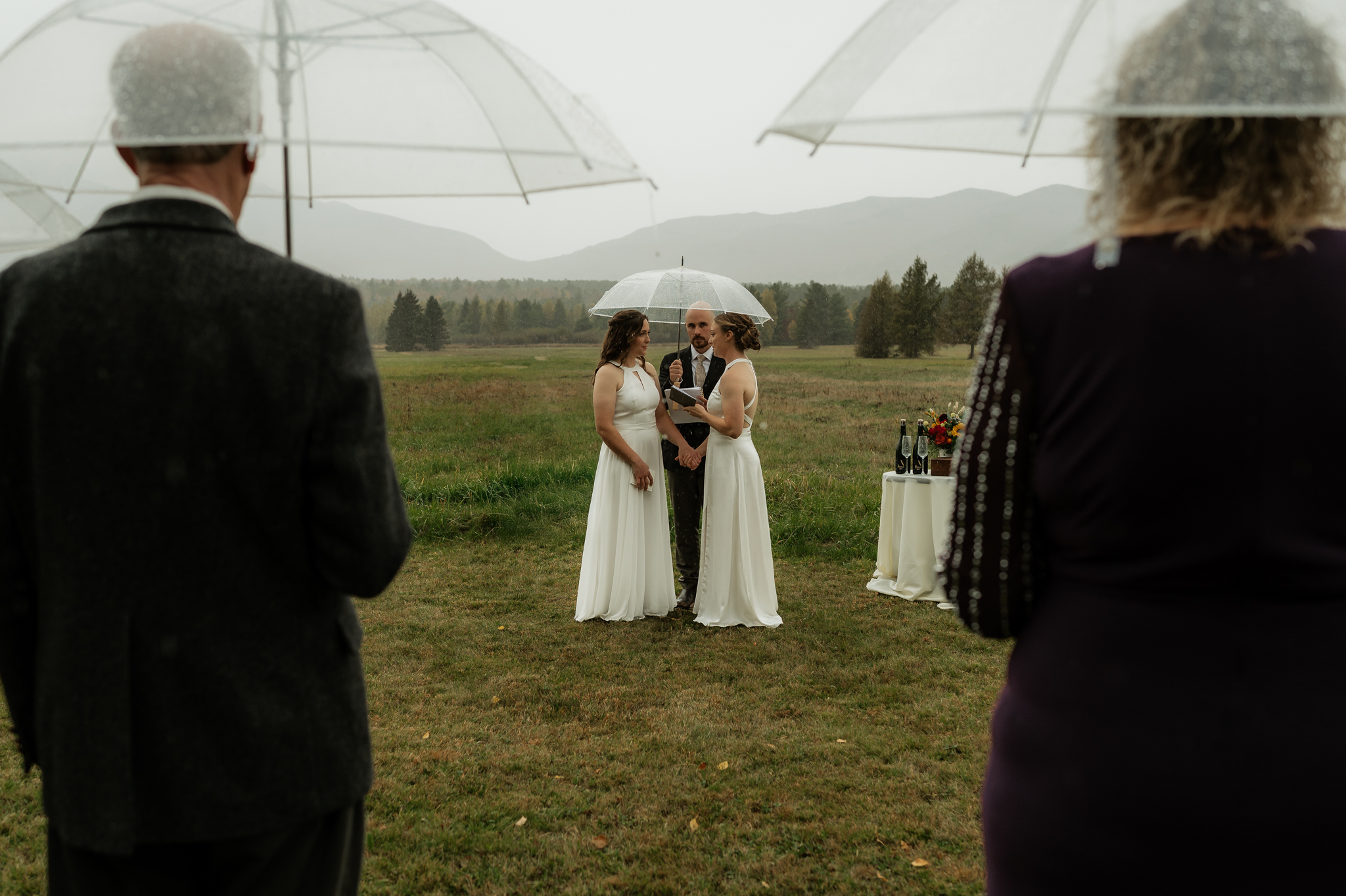A lesbian couple elopes in a field in the Adirondack Mountains in the rain with mountain backdrop.