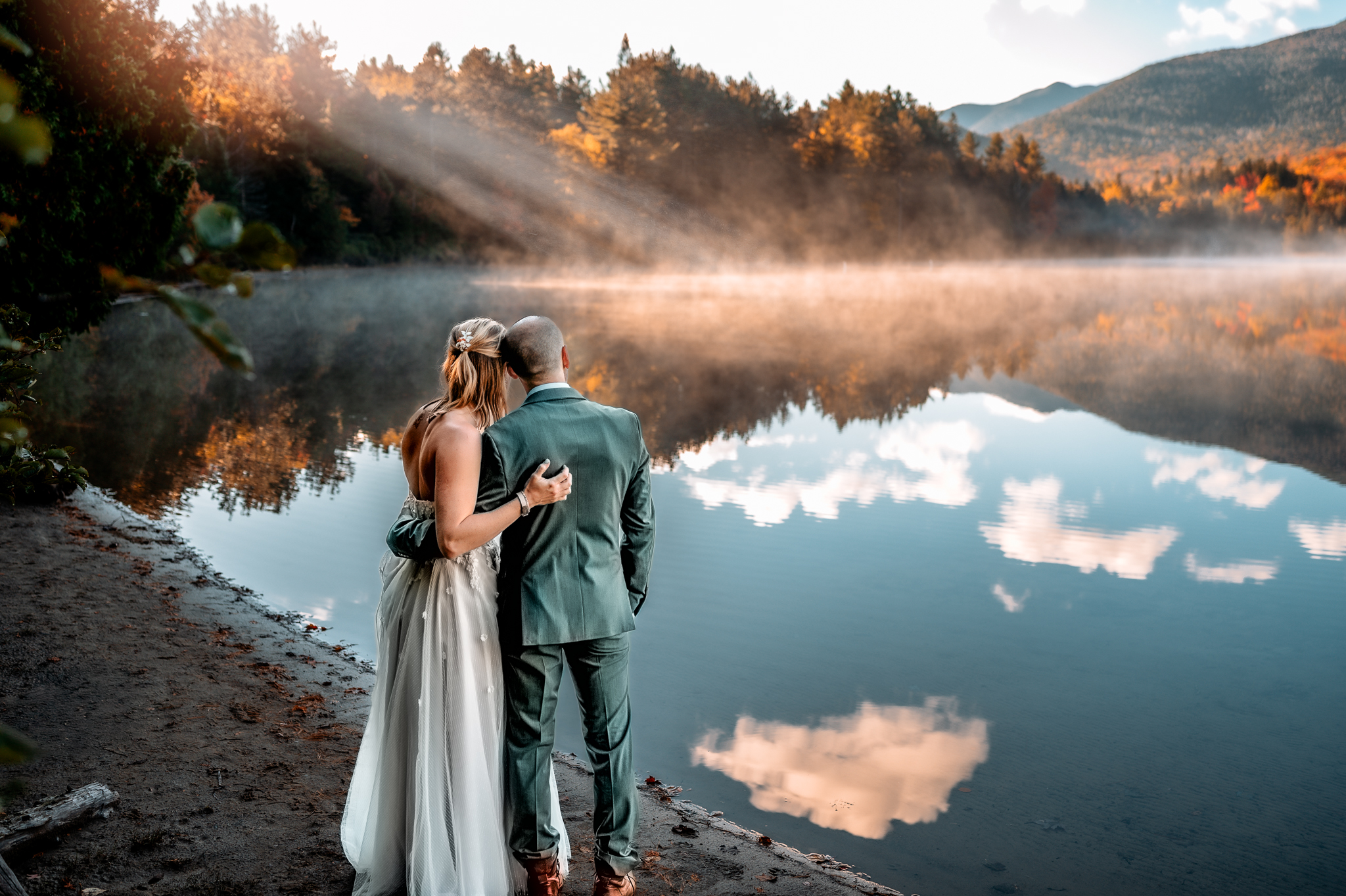 Couple looks out at Heart Lake in the adirondack mountains at sunrise with light rays shining down on the foggy lake and mountains in the background.