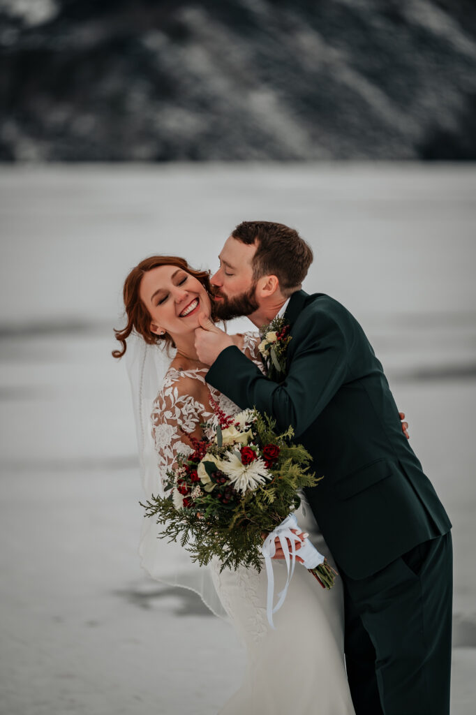 A close up of a bride getting kisses by the groom on a frozen lake after just getting married.