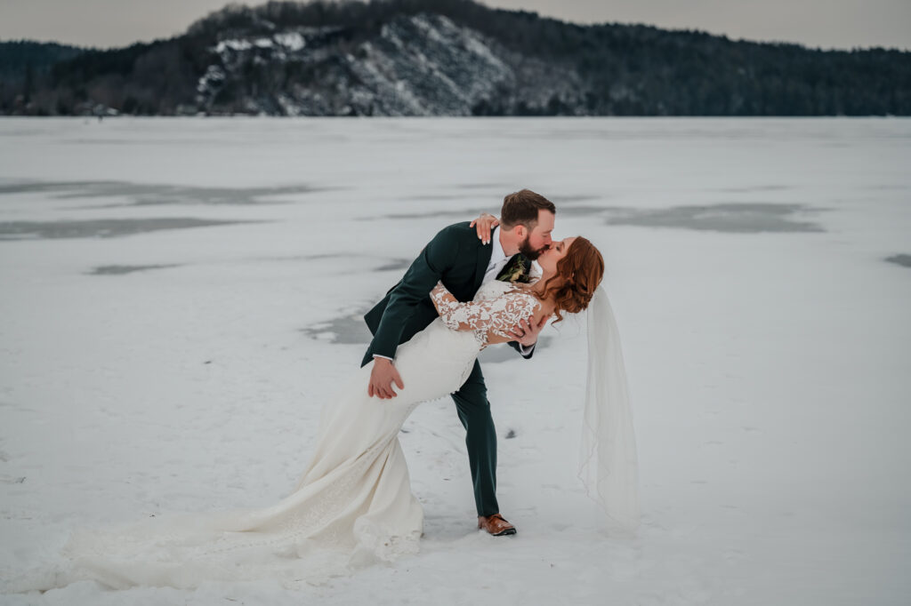 A married couple kisses with a dip on a frozen lake and a mountain hill in the background during winter.