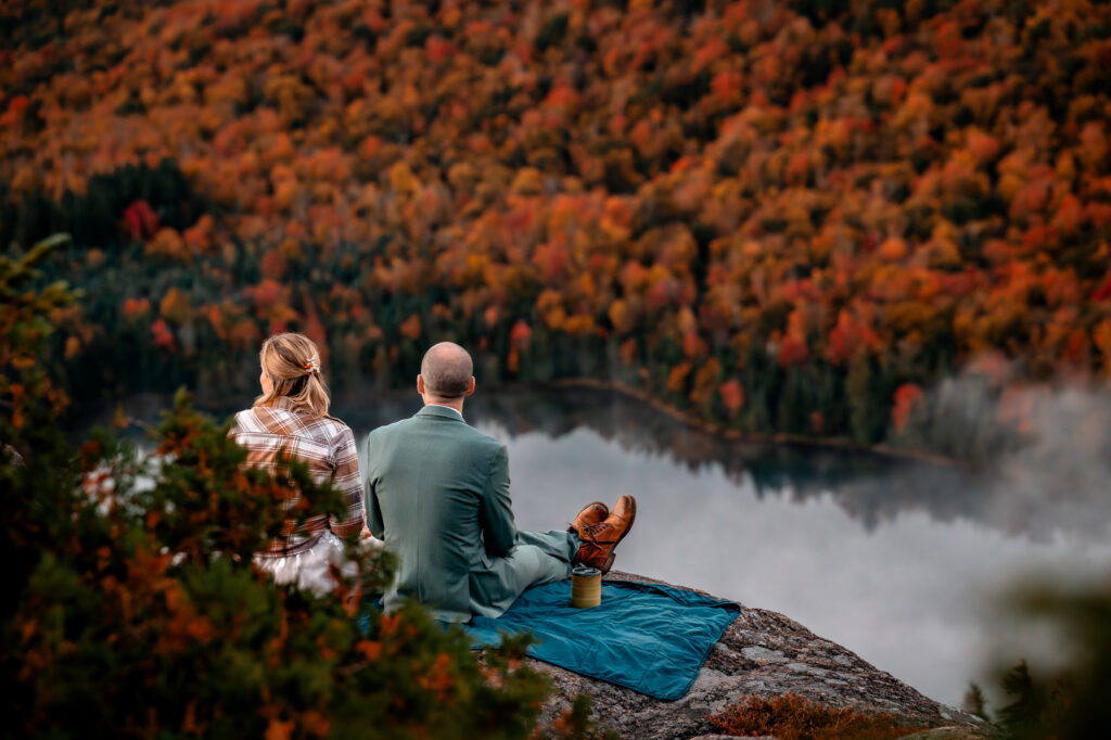 A couple who just eloped sits down on a blue blanket to have coffee on a rock face while looking out at the fall colors on the Mountains overlooking Heart lake which points to how magical it can be to elope in the adirondacks.