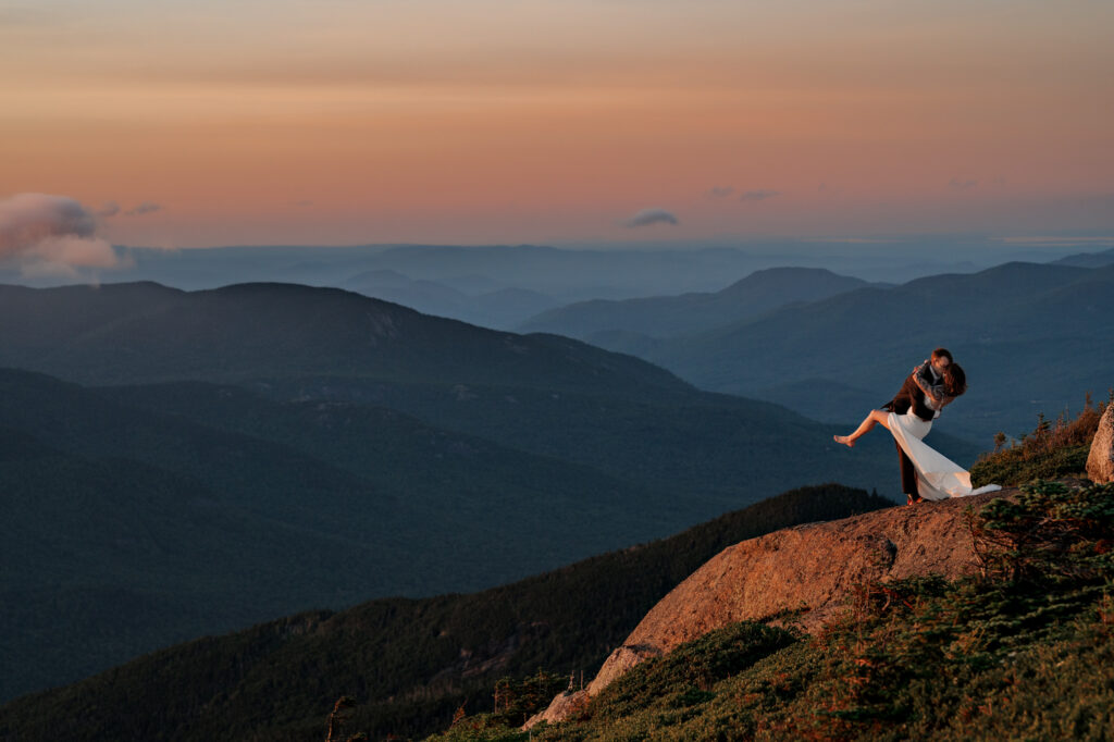 A couple who just eloped embrace at sunset on top of a high peak in the Adirondack Mountains with a mountain backdrop.