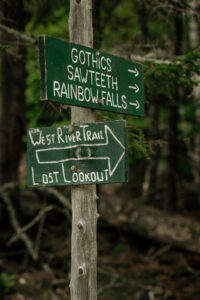 A picture of a trail sign that is green and white that shows the way to gothics mountain high peak in the adirondack mountains.