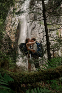 A couple kisses in the blurry background among a large waterfall and pine trees.
