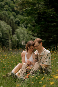 A couple embrace each other and play with their hands as they are sitting in a field of flowers on an Adirondack High peak Hike.
