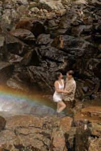 A couple stands on rocks in front of a rainbow at a waterfall in the adirondack mountains.