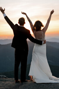 A couple who just eloped on top of a high peak in the adirondack mountains throws up their hands to celebrate completing the hike and getting married at sunset.