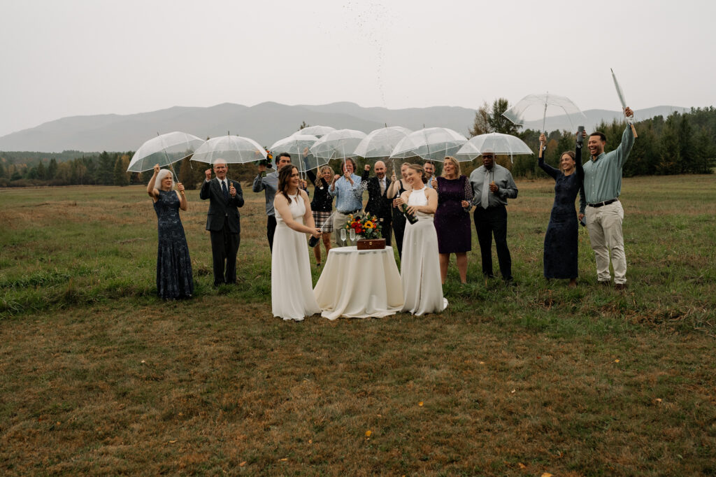 Intimate Adirondack elopement with two brides celebrating with close loved ones under umbrellas in the rain with champagne toast.