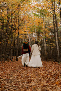 Two brides walk in a forest on a trail with orange and yellow leaves all around them in Autumn with a backpack that has a sign hanging off of it that says married.