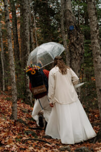Two brides walk through the forest in the fall with an umbrella and a just married sign.