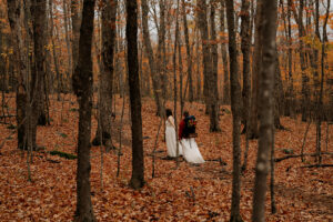 Two brides walk through a forest full of orange and yellow leaves and can be seen in between the trees holding hands.