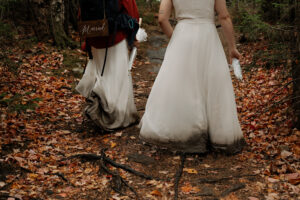 Two brides in white dresses that have dirt and mud all over the bottom of it while walking through a forest floor covered in orange, red and yellow leaves.