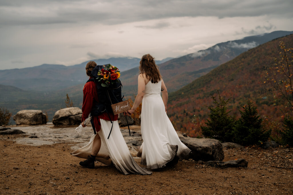 Two brides walking on a windy Adirondack summit in their wedding dresses after their elopement ceremony with muddy dresses as they look out into the distance.