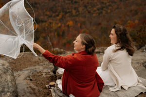 Two brides sitting down to drink hot chocolate on the summit of a mountain when a gust of wind steals their clear umbrella and turns it inside out.