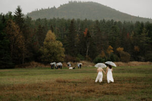 Two brides hold hands walking in the rain with two umbrellas towards their family for an intimate elopement ceremony with a misty mountain backdrop in the Adirondacks.