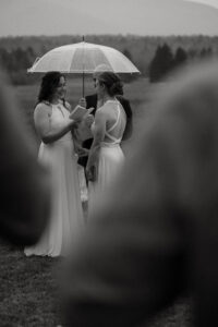 A black and white photo of two brides reading their vows in the rain with an umbrella.