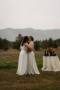 Two brides have their first kiss after saying I DO in a rainy misty field with a mountain backdrop in the Adirondacks.