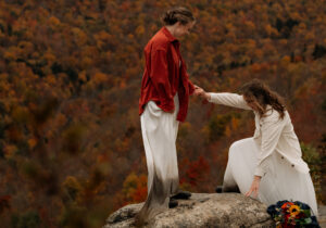 A bride helps her wife up onto a rock in front of a mountain with fall foliage.
