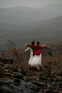 Two brides embracing the wind from a storm moving into the cliff edge they are on in the Adirondack Mountains by putting their arms out like the Titanic.