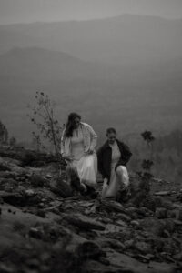 Two brides walk in sync up a rock ledge with mountains behind them.